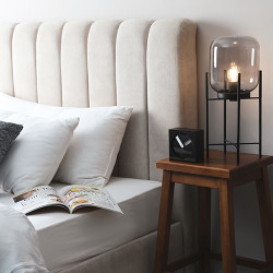 Een bedlampje met een glazen kap en een filigraan zwart metalen onderstel staat op een houten nachtkastje naast een bed met een lichtgekleurd gestoffeerd hoofdeinde